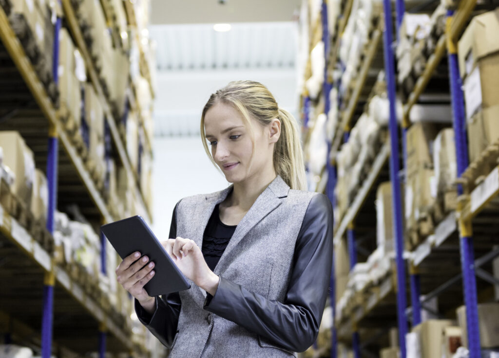 Vertical color image of confident young woman supervisor holding and touching digital tablet in warehouse. Young blond woman standing at distribution warehouse and wearing elegant suit. 实业老板真诚地微笑着看着镜头. Logistic 工作er 工作ing in a large distribution warehouse. Large distribution storage in background with racks and shelfs full of packages, 盒子, 托盘和板条箱准备交付. 物流、货运、海运、收货.